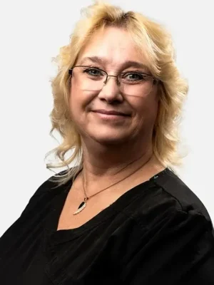 A woman with blonde, wavy hair and glasses is wearing a black top and a necklace, smiling at the camera against a plain white background.